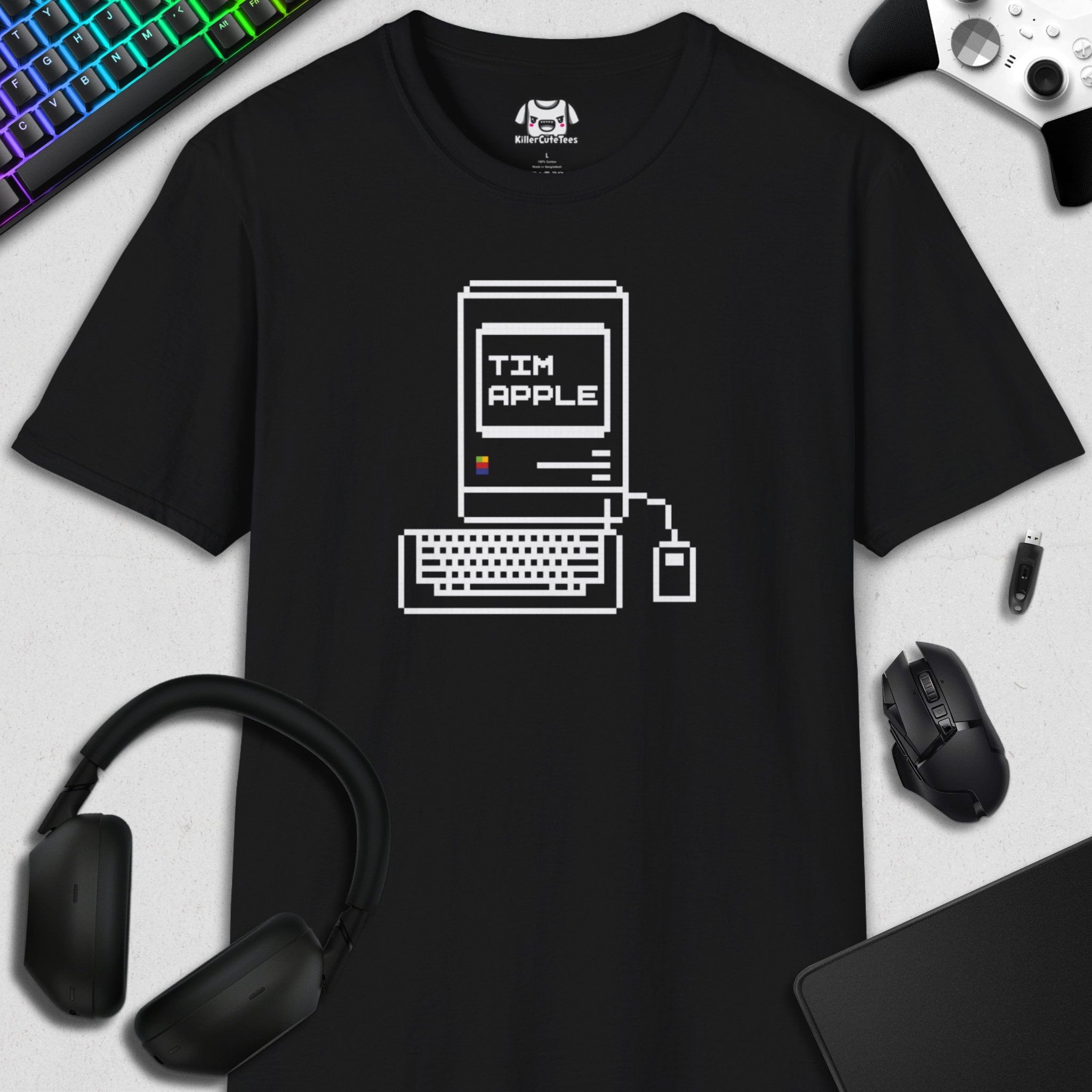 Black t-shirt with computer graphic and 'TIM APPLE' text, surrounded by tech accessories on a light gray background.