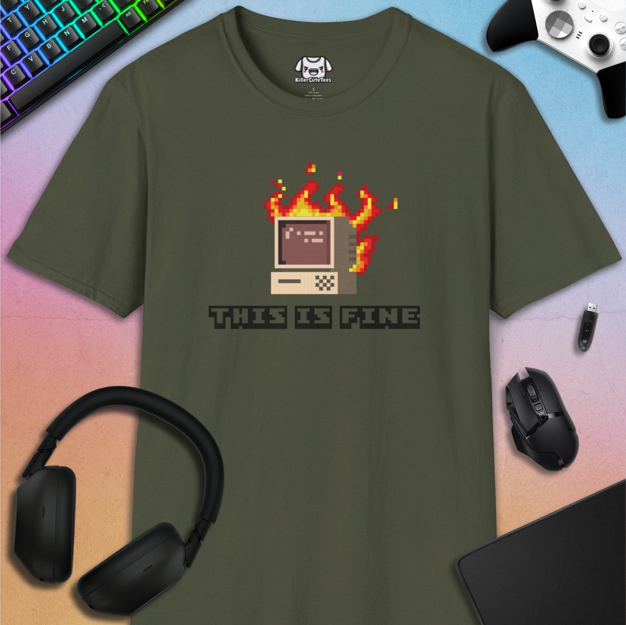Green t-shirt with pixelated computer graphic and 'This is Fine' text, surrounded by gaming accessories on a gradient background.