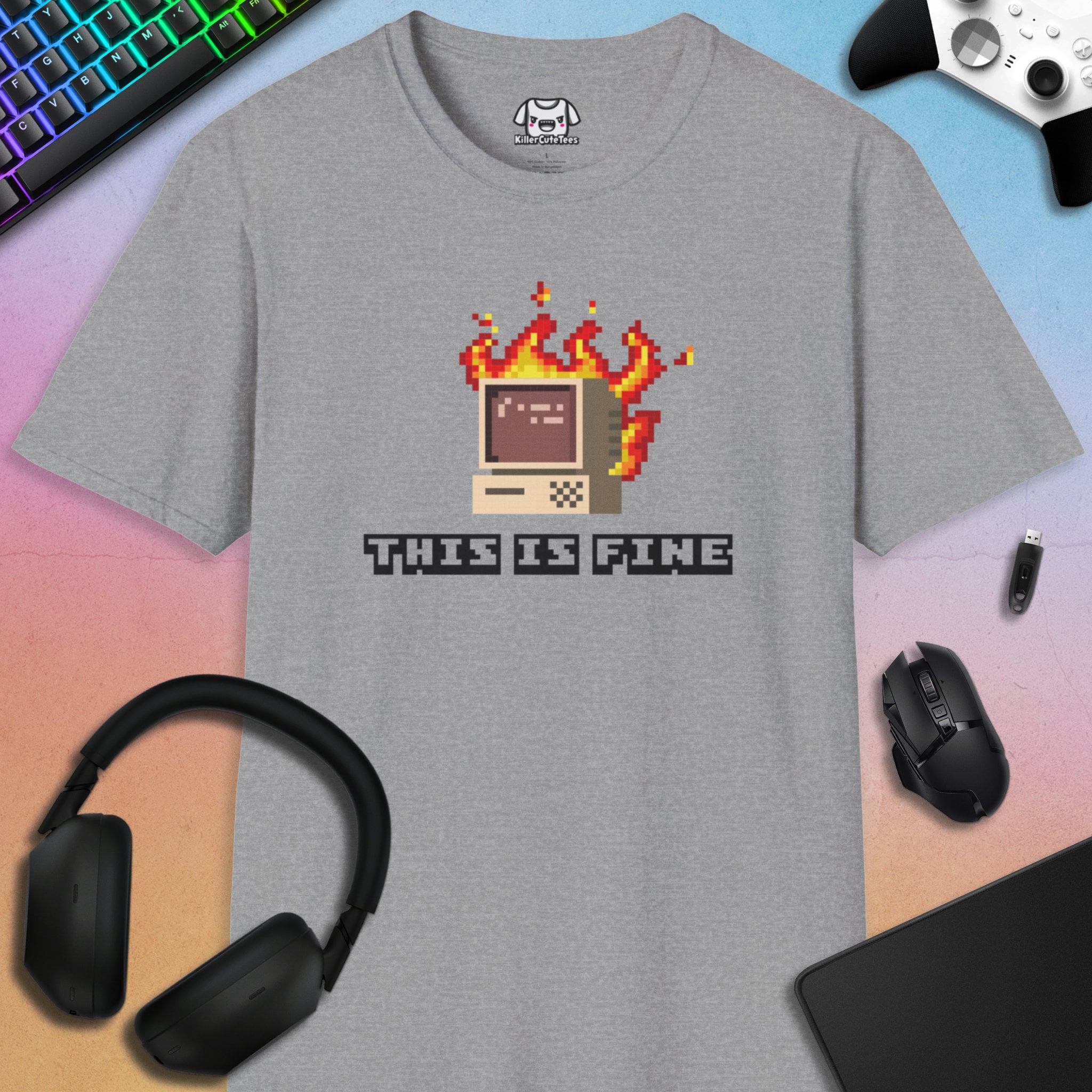 Gray t-shirt with pixel art design and 'This is Fine' text, surrounded by gaming accessories on a gradient background.
